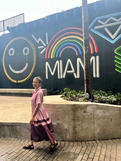 Wided Khadraoui walking past a black wall painted with a neon-style mural reading MIAMI, beside a rainbow, a smiling face, and a diamond.